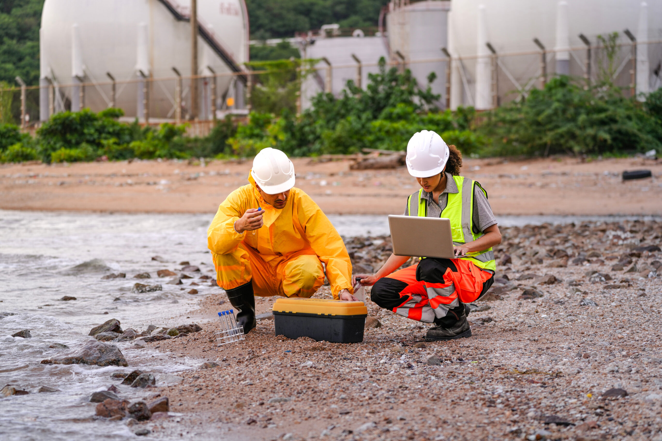Two environmental engineers in safety gear work at a shoreline near gas storage tanks. One collects water samples, while the other records data on a laptop during site inspection. Two environmental engineers in safety gear work at a shoreline near gas storage tanks. One collects water samples, while the other records data on a laptop during site inspection.